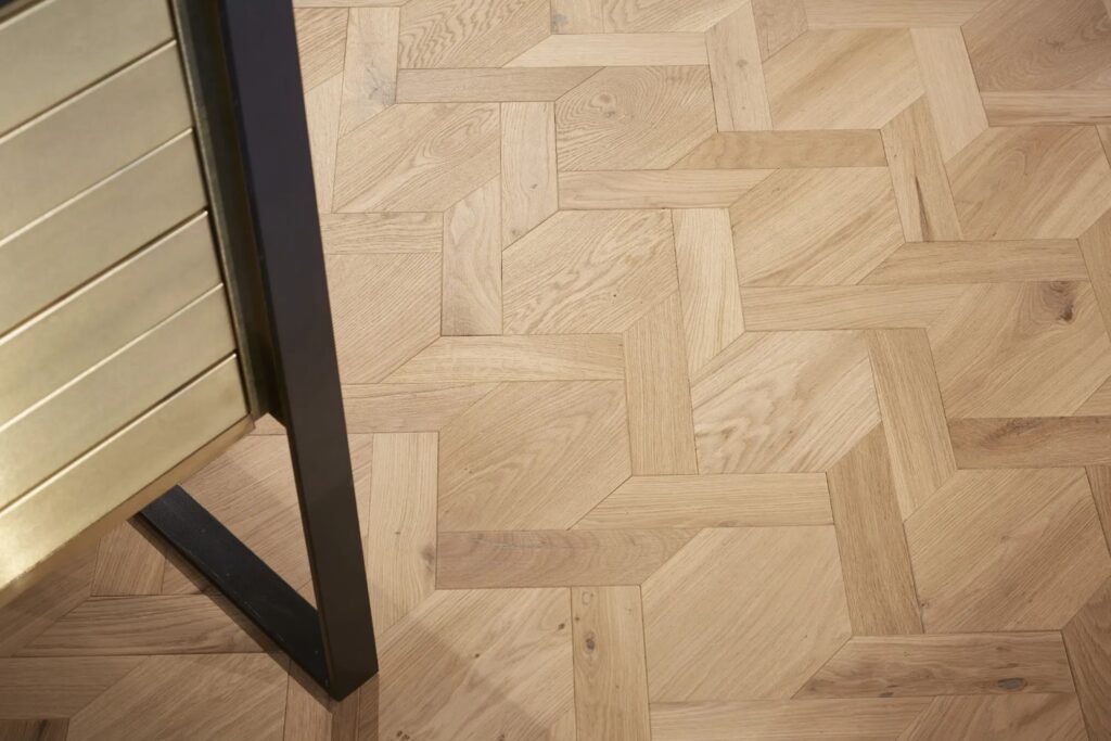 Detailní pohled na luxusní podlahu ve vzoru mozaika v mansion weave stylu z přírodního světlého dřeva, doplněnou o stylový zlatý nábytek a tmavý rám, což vytváří elegantní a sofistikovaný interiérový design.